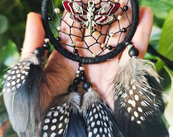 May include: Black dreamcatcher with a silver and red moth pendant. The dreamcatcher features black beads and feathers with white spots. The dreamcatcher is held in a hand.