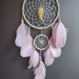 Op de afbeelding: Een beige droomvanger met een wit web en turquoise kralen. De droomvanger heeft een gouden veerbedel en roze veren die eronder hangen.