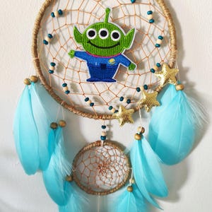 Puede incluir: Un atrapasueños hecho a mano con un aplique de personaje alienígena verde. El atrapasueños tiene un diseño de telaraña tejida, adornado con plumas azul claro, cuentas de madera y detalles de estrellas doradas. El anillo exterior está envuelto en hilo de color natural.