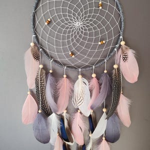 Peut inclure: Un attrape-rêves gris et blanc avec des plumes roses, blanches et grises. L'attrape-rêves est orné de perles en bois et d'un cercle en macramé gris.