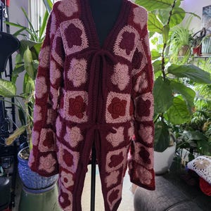 Puede incluir: Un cárdigan hecho a mano, abierto por delante, en tonos burdeos y rosa claro. El diseño de ganchillo presenta motivos cuadrados con patrones florales. El cárdigan tiene mangas largas y un cierre de lazo en la cintura.