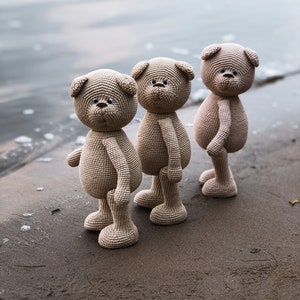 May include: Three brown crocheted teddy bears standing on a sandy beach. The bears have black eyes and noses and are facing forward.