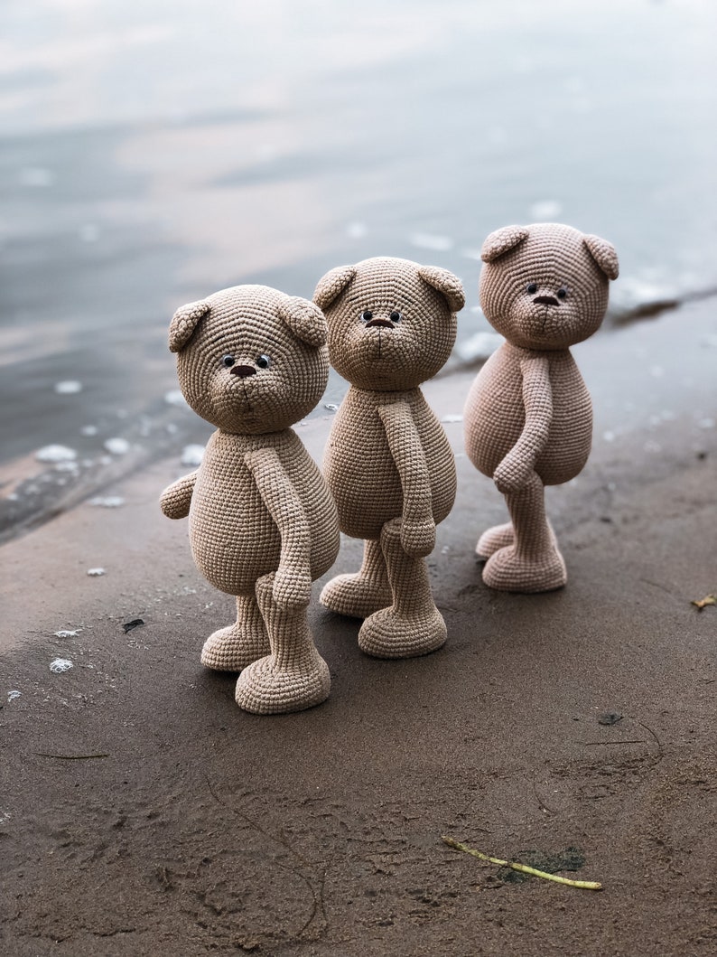 May include: Three brown crocheted teddy bears standing on a sandy beach. The bears have black eyes and noses and are facing forward.