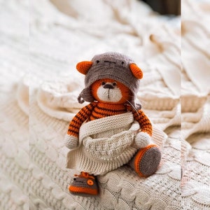Puede incluir: Una muñeca de tigre hecha a mano de ganchillo naranja y marrón que lleva un sombrero gris con orejas naranjas, una bufanda color crema y zapatos a juego. El tigre tiene rayas negras y está sentado sobre una manta de punto color crema.