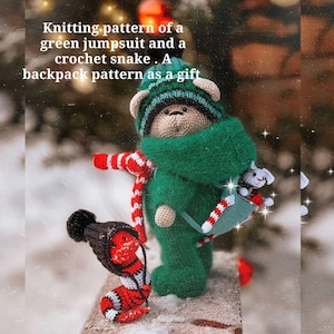 以下が含まれることがあります： 緑色のつなぎと赤と白のストライプのストールを着用した、かぎ針編みで作られたテディベア。クマは緑色のバックパックに、かぎ針編みで作られたヘビを入れています。画像には「Knitting pattern of a green jumpsuit and a crochet snake. A backpack pattern as a gift」というテキストが表示されています。