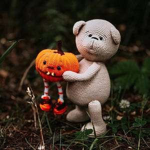 May include: A crocheted plush toy bear holding a crocheted pumpkin with a jack-o'-lantern face. The bear and pumpkin are standing on a bed of brown pine needles and green grass.