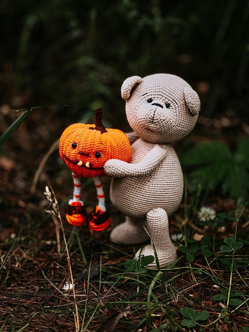 May include: A crocheted plush toy bear holding a crocheted pumpkin with a jack-o'-lantern face. The bear and pumpkin are standing on a bed of brown pine needles and green grass.