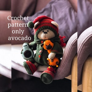 Puede incluir: Patrón de crochet para un oso de peluche verde aguacate que lleva un gorro rojo y verde y sostiene un pequeño animal de crochet naranja y marrón. El texto "Crochet pattern only avocado" es visible en la imagen.