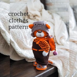 Op de afbeelding: Gehaakte kat die een bruin en oranje outfit draagt met een grijze hoed en een bruine rugzak. De kat staat op een houten oppervlak. crochet clothes pattern