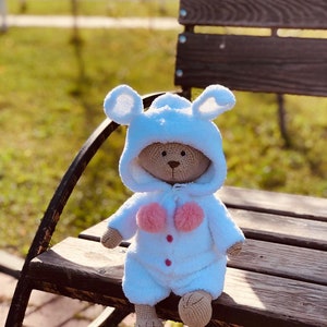 Puede incluir: Un oso de peluche blanco de crochet que lleva un mono azul con capucha y pompones rosas. El oso está sentado en un banco de madera.