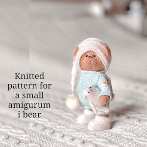 May include: A small, crocheted amigurumi bear wearing a light blue sweater, white hat, and boots, holding a white stuffed animal. The text "Knitted pattern for a small amigurumi i bear" is on the left.