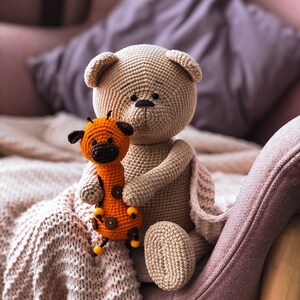 May include: A crocheted beige teddy bear holding a crocheted orange giraffe toy. The toys are sitting on a pink armchair with a white knitted blanket.