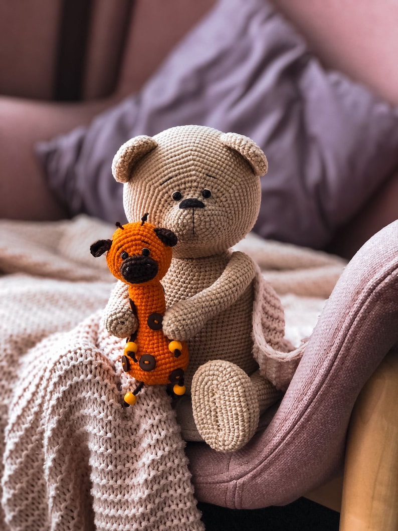 May include: A crocheted beige teddy bear holding a crocheted orange giraffe toy. The toys are sitting on a pink armchair with a white knitted blanket.