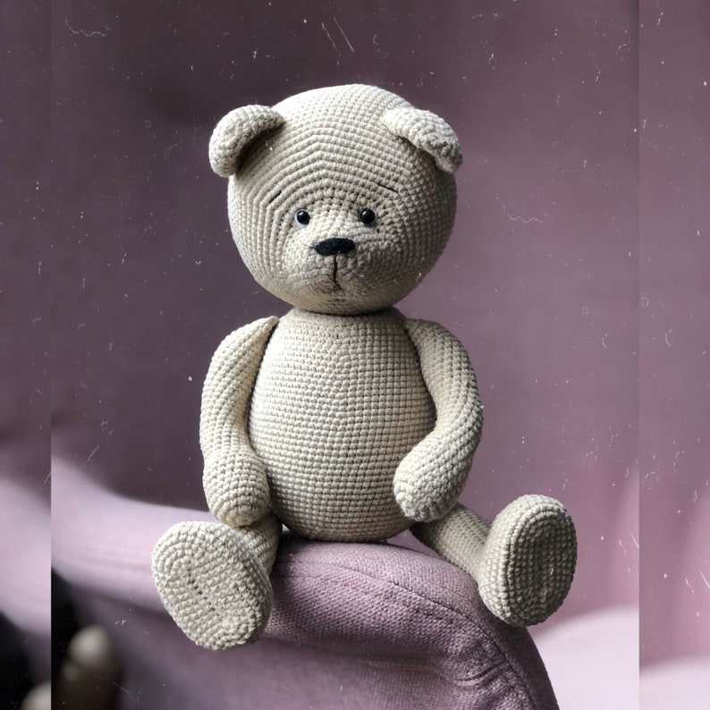 May include: A crocheted beige teddy bear with black eyes and a black nose sits on a pink chair.