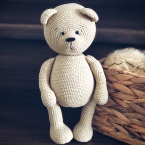 May include: A crocheted beige teddy bear with black eyes and a black nose. The bear is standing on a wooden surface with a wicker basket behind it.