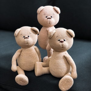 May include: Three crocheted teddy bears sitting on a black couch. The bears are beige and have brown eyes and noses.