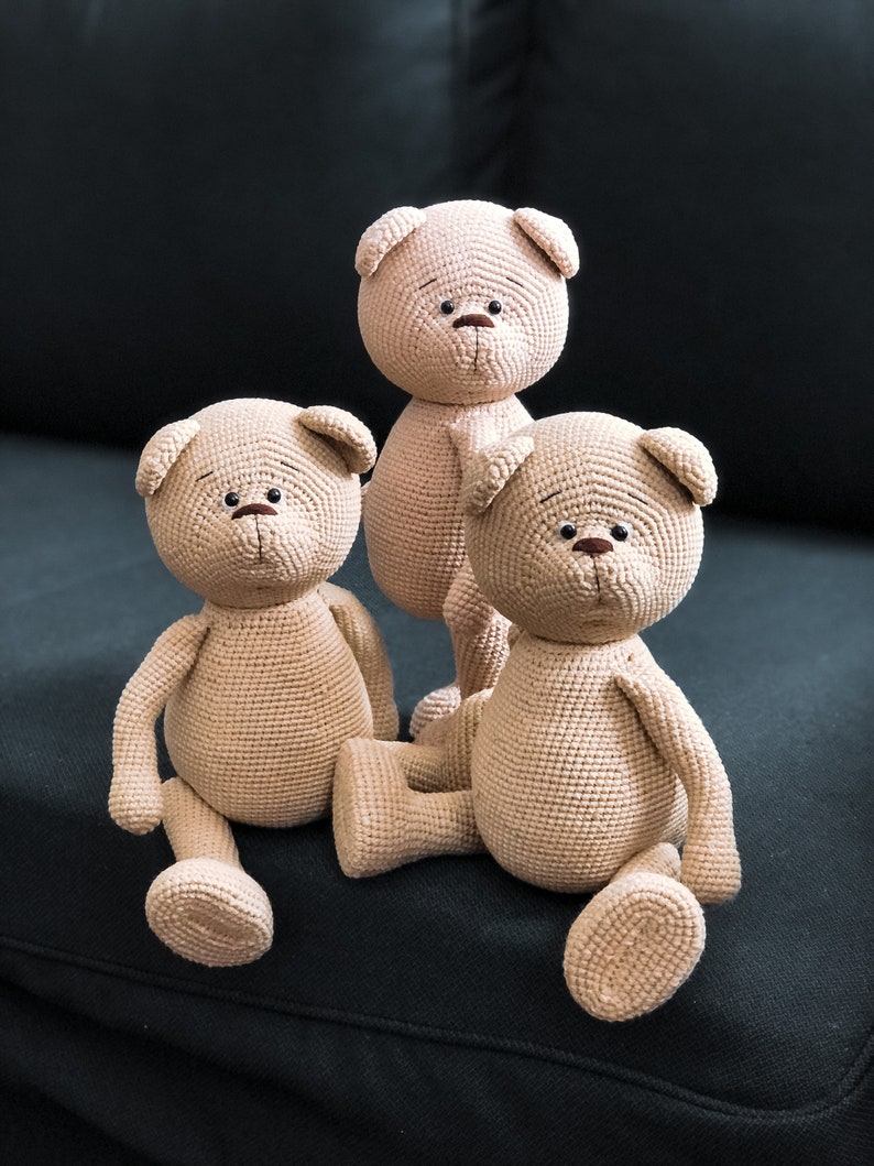 May include: Three crocheted teddy bears sitting on a black couch. The bears are beige and have brown eyes and noses.