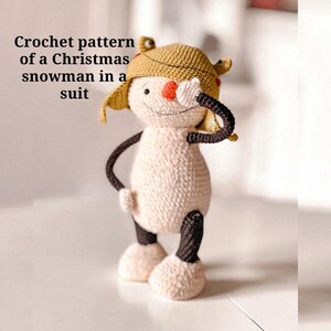 May include: A crocheted snowman in a suit, wearing a tan hat with ear flaps. The snowman has a red nose, black arms, and black and white legs. The text "Crochet pattern of a Christmas snowman in a suit" is in the image.