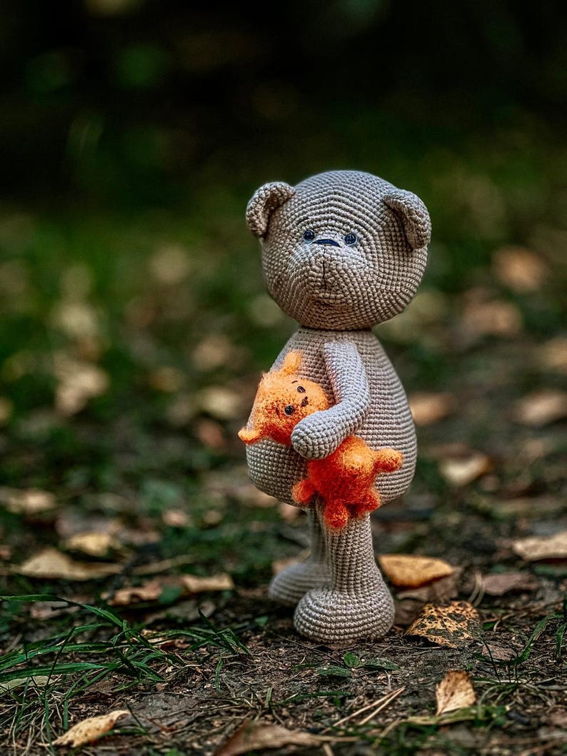 May include: A gray crocheted teddy bear holding a smaller orange crocheted teddy bear. Both bears have black eyes and a stitched smile.