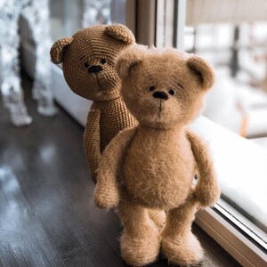 May include: Two brown crocheted teddy bears standing next to a window. The bear in the front is fuzzy and the bear in the back is smooth.