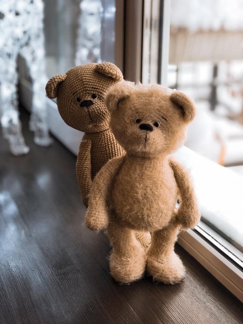 May include: Two brown crocheted teddy bears standing next to a window. The bear in the front is fuzzy and the bear in the back is smooth.