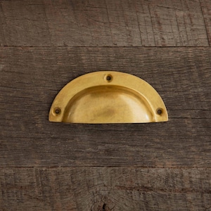Könnte beinhalten: Ein messingfarbener, halbmondförmiger Schubladengriff mit drei Schraubenlöchern. Die Hardware befindet sich auf einer verwitterten, braunen Holzoberfläche. Der Griff hat ein gebogenes, vertieftes Design für einfaches Greifen.