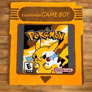 May include: A yellow and brown rug shaped like a Game Boy cartridge with a pixelated image of Pikachu from the Pokemon video game series. The rug has the text "Nintendo GAME BOY" and "Nintendo" on it.