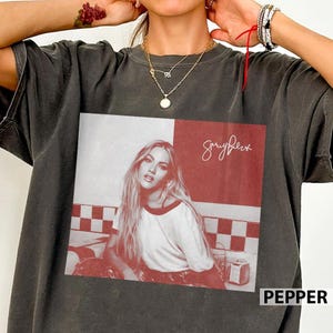 May include: A charcoal gray short-sleeve t-shirt featuring a square graphic print. The print shows a black and white portrait of a woman with long hair wearing a t-shirt, set against a red and white checkered background with the script text "Garryfulez".
