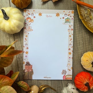 Peut inclure: Un bloc-notes blanc avec une bordure sur le thème de l'automne représentant des citrouilles, des feuilles, des bougies et une cheminée. Le bloc-notes porte le mot "Notes" en haut et le mot "Amourette" en bas.