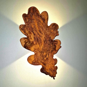 May include: A wall-mounted light fitting shaped like an oak leaf with a textured, rusted brown finish. The fitting emits a soft, warm glow from behind its edges, creating a distinct light pattern on the wall surface behind the object.