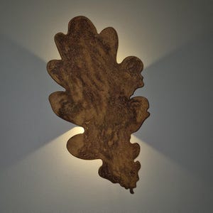 Op de afbeelding: Een decoratieve wandlamp in de vorm van een eikenblad. Het blad heeft een gevlekte bruine kleur met een textuuroppervlak. Twee lampen verlichten de lamp en werpen schaduwen op de muur. Ongeveer 50 cm hoog.