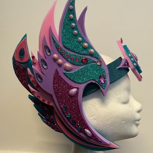 May include: A colorful, layered headpiece with a fantasy theme. The headpiece features shades of pink, purple, and teal, with glitter accents and decorative jewels. The design includes curved, wing-like shapes and is mounted on a dark teal headband.