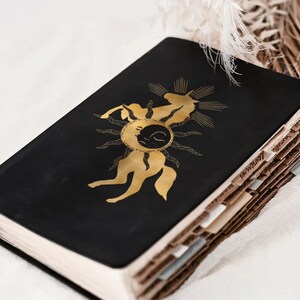 K&ouml;nnte beinhalten: Ein schwarzes, ledergebundenes Tagebuch mit einem goldenen Einhorn-Design auf dem Einband. Das Einhorn ist von einem Sonnenstrahl und einem Halbmond umgeben. Das Tagebuch ist geschlossen und liegt auf einer wei&szlig;en Oberfl&auml;che.