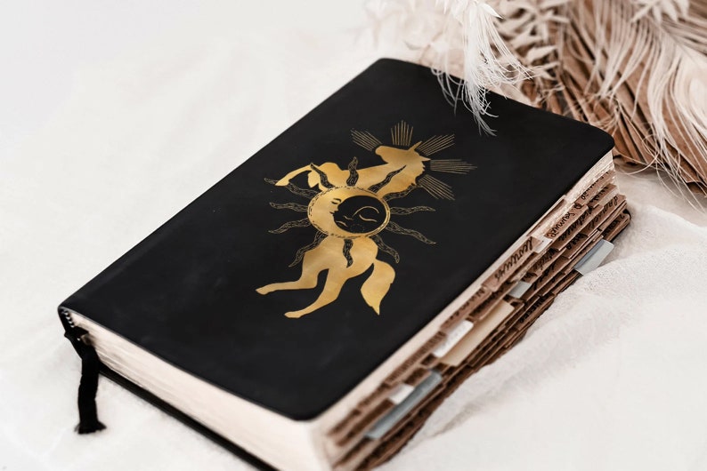 K&ouml;nnte beinhalten: Ein schwarzes, ledergebundenes Tagebuch mit einem goldenen Einhorn-Design auf dem Einband. Das Einhorn ist von einem Sonnenstrahl und einem Halbmond umgeben. Das Tagebuch ist geschlossen und liegt auf einer wei&szlig;en Oberfl&auml;che.