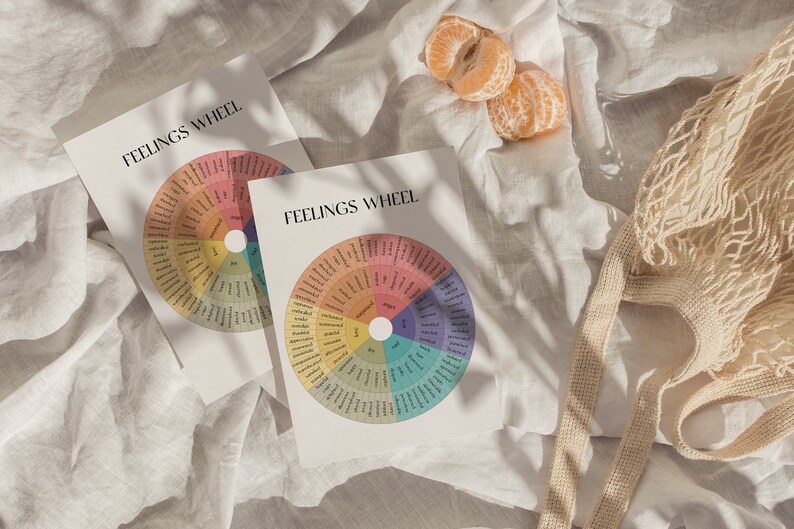 May include: Two printable "Feelings Wheel" charts on white paper with a colorful wheel of emotions. The charts are on a white linen fabric background with a beige mesh bag and two halved oranges.