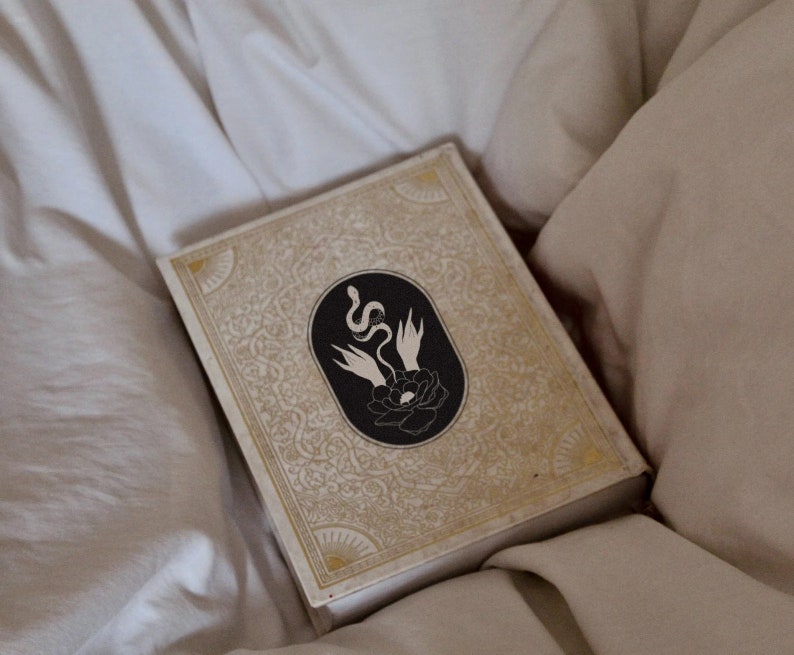 K&ouml;nnte beinhalten: Ein geschlossenes Buch mit einem gold- und wei&szlig; gemusterten Einband. Der Einband zeigt eine schwarz-wei&szlig;e Illustration einer Schlange, die sich um eine Blume windet.