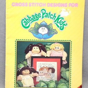 May include: A cross stitch pattern booklet featuring Cabbage Patch Kids dolls. The cover features three dolls with blonde, brown, and dark brown hair. The title of the booklet is "Cabbage Patch Kids" and the text "All Babies are Special but Are Found in a Cabbage Patch" is included in a cross stitch design.