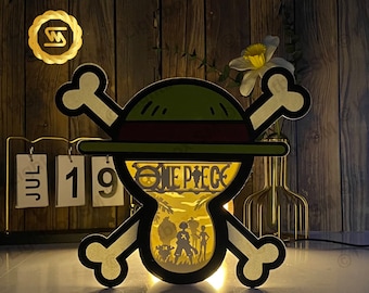 May include: A wooden lamp shaped like the One Piece logo, with a straw hat and the words "One Piece" on the front. The lamp is lit and casting a warm glow.