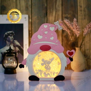 May include: A pink wooden gnome night light with a white heart and a Mickey Mouse and Minnie Mouse silhouette inside. The gnome has a pink hat with white hearts and a bow.
