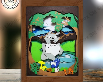 May include: A papercraft scene featuring a grey and white Totoro character hanging from a tree branch in a woodland setting. The scene includes green trees, blue sky, brown mountains, and a blue river. There are also small yellow and orange mushrooms in the foreground.