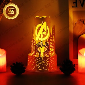 May include: A white paper lamp with a cut-out design of two superheroes fighting in front of a cityscape. The Avengers logo is visible in the background. The lamp is lit from the inside, casting a warm glow.