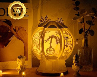 May include: A white papercraft lamp shaped like a sphere with cutouts of the Grinch and the words "The Grinch" and "Ho Ho Ho". The lamp is lit from within, casting a warm glow.