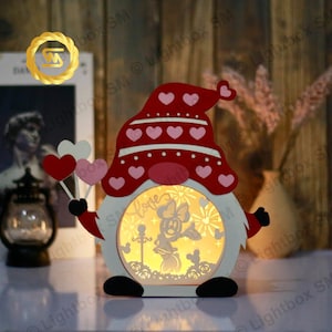 May include: A white wooden gnome figurine with a red hat decorated with pink hearts. The gnome is holding a balloon with a heart shape. The gnome's belly is a clear window that reveals a light-up scene of a cartoon character with a bow and fireworks. The scene says "Love" in a decorative font.