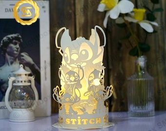 Pode incluir: Uma lâmpada de papel branco iluminada com um design recortado de Stitch do filme da Disney Lilo & Stitch. A lâmpada está acesa e a luz brilha através dos recortes. A lâmpada está sobre uma superfície branca com um livro e um vaso de vidro com flores no fundo.