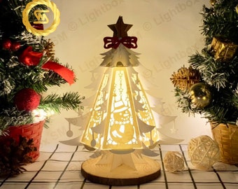 May include: A white wooden Christmas tree with a snowman design and a red bow on top. The tree is illuminated from within, casting a warm glow. The tree is sitting on a wooden base and is surrounded by greenery.