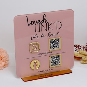 Könnte beinhalten: Ein rosa Acrylschild mit goldener Beschriftung, auf dem "Loved & Link'd Let's be Social" steht. Das Schild hat vier goldene Symbole, darunter eine Kamera, ein Facebook-Logo und zwei QR-Codes. Das Schild steht auf einem goldenen Ständer.