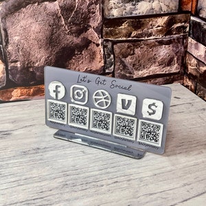 May include: A grey acrylic sign with the text "Let's Get Social" and icons for Facebook, Instagram, Twitter, Vimeo, and Venmo. Each icon has a corresponding QR code underneath.