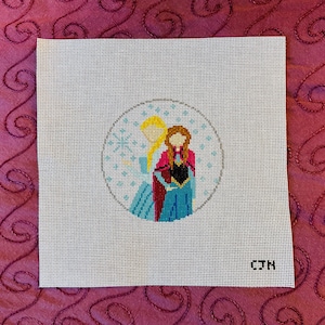 May include: A white canvas with a cross-stitch design of two girls, one with blonde hair and one with red hair, standing inside a circle. The circle has a snowflake design. The canvas has the text "CJN" in the bottom right corner.
