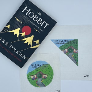 May include: Two needlepoint canvases with a quote from The Hobbit, "Not all those who wander are lost." One canvas is a circle with a green and brown hobbit hole, the other is a triangle with a blue sky and a green hill.