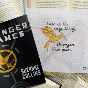 May include: A copy of "The Hunger Games" book by Suzanne Collins is next to a needlepoint with a gold bird and the text "hope is the only thing stronger than fear". The book cover is black with a gold bird.